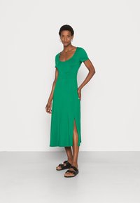 Vestido verde até ao joelho em tecido suave com decote redondo, mangas curtas e fendas laterais; combinado com sandálias pretas.