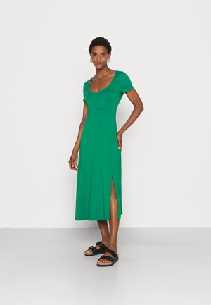 Vestido verde até ao joelho em tecido suave com decote redondo, mangas curtas e fendas laterais; combinado com sandálias pretas.