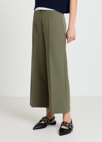 Olivgrüne, weit geschnittene Hose aus glattem Material mit vorderen Falten und legerer Passform. Getragen mit schwarzen Slingback-Schuhen mit spitz zulaufender Spitze.