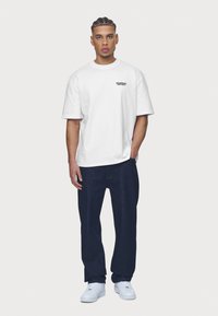 White cotton t-shirt with short sleeves, featuring a small black logo on the left chest; paired with dark blue denim pants and white sneakers.