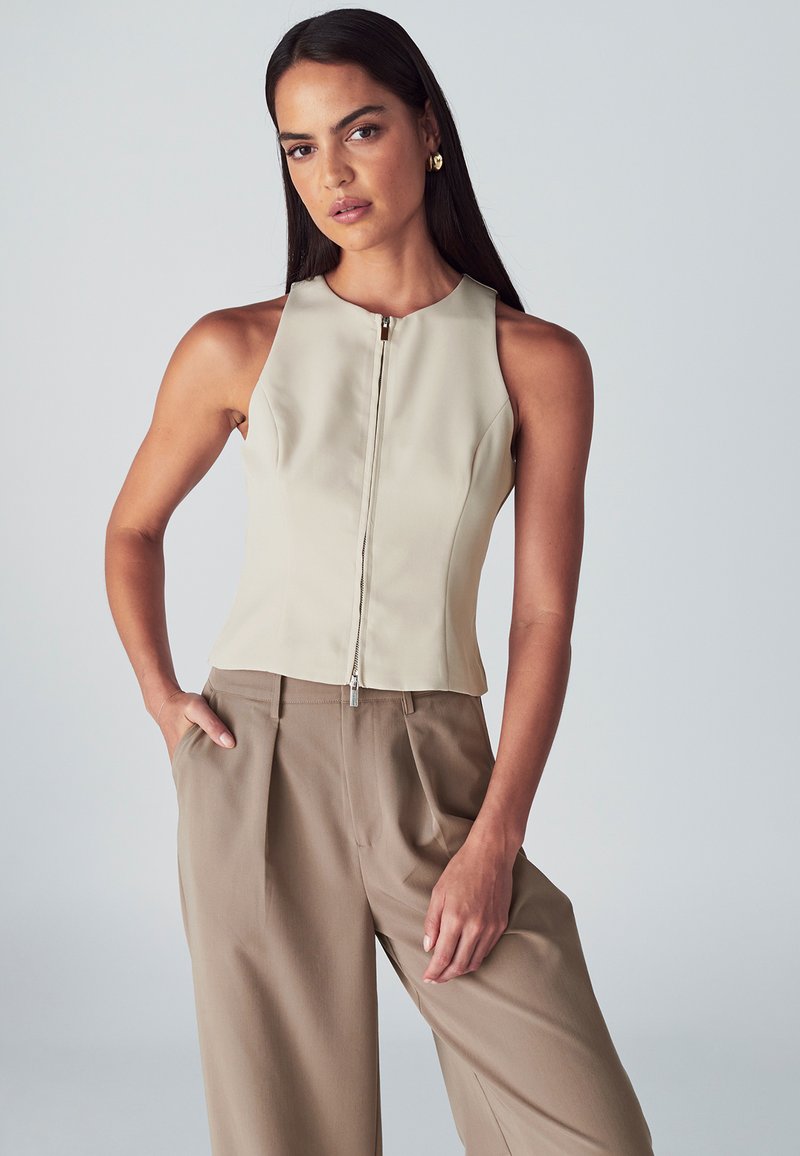 Top beige senza maniche con collo alto e zip frontale, abbinato a pantaloni khaki larghi a vita alta con tasche laterali.