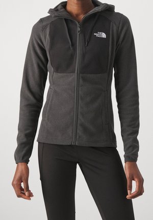 Persoon die een donkergrijze zip-up capuchonjas met The North Face-logo op de borst en zwarte nauwsluitende broek draagt tegen een effen achtergrond.