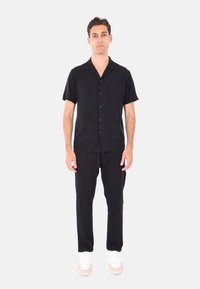 Camicia nera a maniche corte con bottoni e tasca, abbinata a pantaloni neri. Il modello indossa scarpe da ginnastica bianche. Design semplice, tessuto leggero.