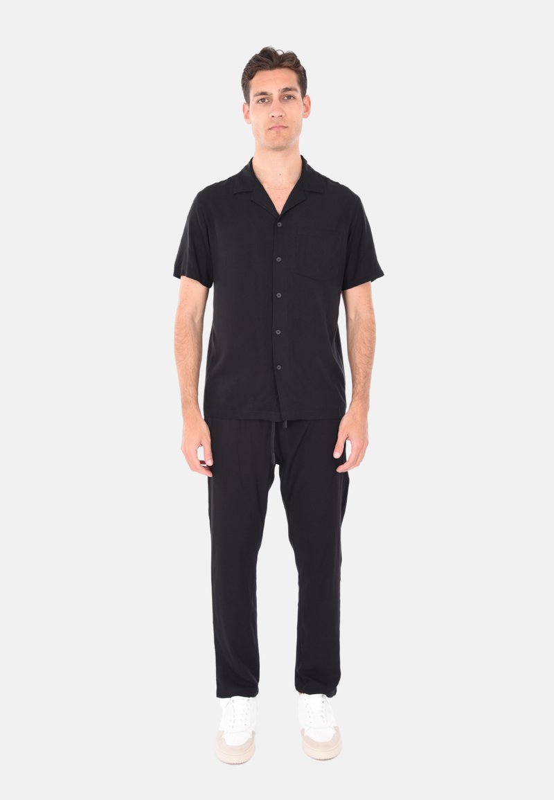 Camicia nera a maniche corte con bottoni e tasca, abbinata a pantaloni neri. Il modello indossa scarpe da ginnastica bianche. Design semplice, tessuto leggero.