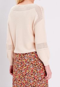 Světlý béžový pletený svetr s texturovaným vzorem a otevřenými vzory na ramenou, doplněný barevnou sukní s potiskem.