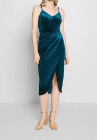 Robe midi en velours bleu sarcelle avec un corsage ajusté, un décolleté en V et un détail noué en forme de drapé, comprenant une fente haute sur la cuisse et des bretelles réglables.