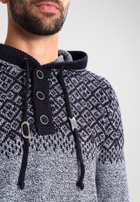 Marineblauer Strick-Hoodie mit geometrischem Muster, Knopfleiste am Hals und Kordelzug. Strukturierter Stoff mit kontrastierenden Farben.