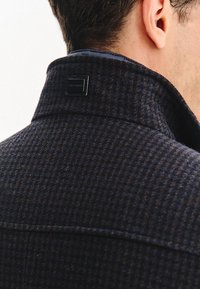 Tekstylna marynarka w kolorze granatowym i brązowym w wzór houndstooth z kołnierzem, z czarną markową metką z tyłu, blisko dekoltu.