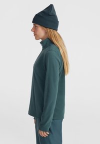 Fleece svetr v tmavě modré barvě s vysokým límcem, přiléhavými manžetami a kapsou na prsou. Kombinováno s tmavě modrými kalhotami a odpovídající čepicí.