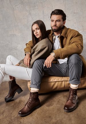 Bruine leren veterschoenen met rubberen zolen, gecombineerd met grijze broek en een zacht beige trui. Gladde textuur met een contrasterend paneeldetail.