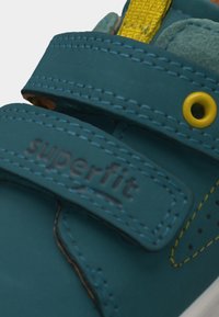 Κοντινό πλάνο παιδικού παπουτσιού σε δαντελένιο μπλε χρώμα με κίτρινες λεπτομέρειες και λουράκια με βέλκρο, με την ένδειξη "superfit" χαραγμένη στον πάνω ιμάντα.