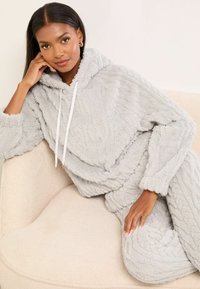 Set da lounge grigio, morbido e con cappuccio, caratterizzato da un motivo testurizzato. Presenta un cappuccio con coulisse e maniche lunghe accoglienti, indossato mentre si è seduti su una morbida sedia beige.