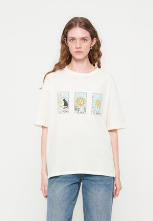 Νεαρή γυναίκα με μακριά κυματιστά μαλλιά φοράει λευκό φαρδύ t-shirt με σχέδια από κάρτες ταρώ: Η Σελήνη, Ο Ήλιος και Το Αστέρι, σε συνδυασμό με μπλε τζιν.