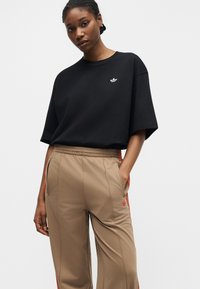 Femme debout portant un T-shirt noir ample avec un petit logo blanc et un pantalon de survêtement beige avec des bandes rouges et un logo, les mains dans les poches.
