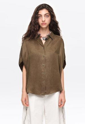 Joven mujer con cabello largo y rizado que lleva una blusa verde oliva abotonada y pantalones blancos con cremalleras laterales, posando frente a un fondo liso.