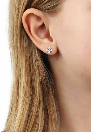 Primo piano dell'orecchio di una persona con capelli biondi lisci che indossa un piccolo orecchino a bottone rotondo in argento con una pietra preziosa trasparente.