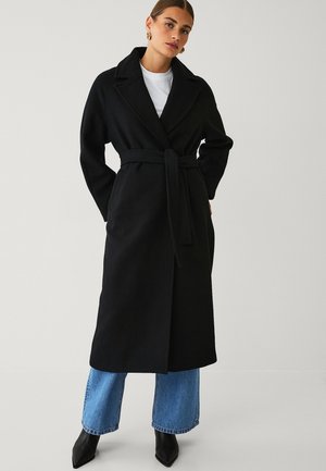 Kvinde iført en lang sort kåbe med bælte over en hvid skjorte, blå jeans og sorte spidse støvler, stående mod en ensfarvet lys baggrund.