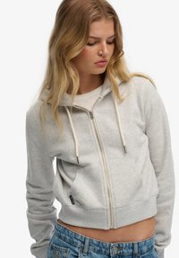 Szara krótka bluza z zamkiem wykonana z miękkiego materiału, z kapturem, sznurkami, przednimi kieszeniami i ściągaczami przy mankietach. Nosi się ją na jasnoniebieskich dżinsach.