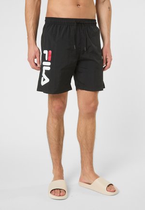 Mężczyzna w czarnych kąpielówkach FILA i beżowych klapkach stoi na jednolitym jasnym tle.