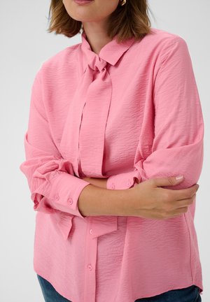 Femme portant un chemisier rose texturé à manches longues avec un col noué, tenant son bras gauche croisé sur son corps, visible des épaules jusqu'aux hanches.