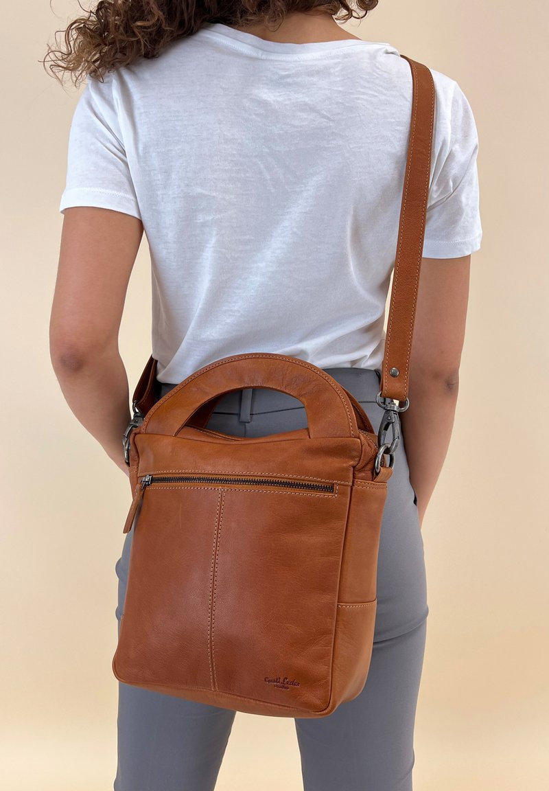 Brązowa skórzana torebka na ramię z zaokrągloną rączką, regulowanym paskiem, przednią kieszenią na zamek błyskawiczny oraz detalami w szwie. Gładka tekstura, minimalistyczny design.