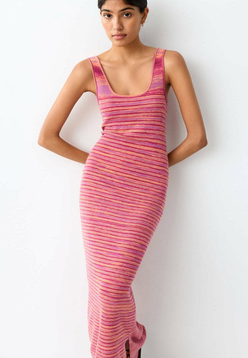 Jeune femme debout contre un mur blanc, portant une robe en maille sans manches, moulante, à rayures roses et orange, avec une fente près de l'ourlet.