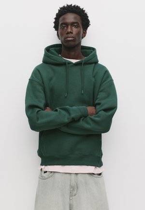 Sweat-shirt à capuche vert en coton avec une coupe décontractée, doté d'une poche kangourou et de poignets ribbed. Porté par-dessus une chemise rose clair.