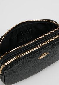 Musta nahkapussi kultaisella vetoketjulla, jossa on teksturoidun pinnan ja logoyksityiskohta edessä. Sisäpuoli on vuorattu mustalla kankaalla.