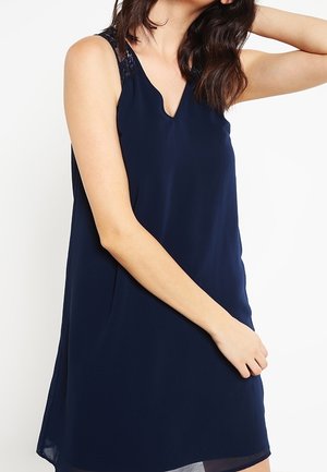 Kvinde iført en ærmeløs mørkeblå kjole med blonde detaljer på skuldrene, poserer med den ene arm løftet og den anden afslappet.