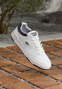Adidași albaștri din piele cu accente negre, vârf perforat, șireturi plate și un logo verde de crocodil pe lateral. Talpă din cauciuc și guler căptușit.