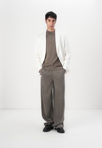 Blazer bianco su un dolcevita marrone e pantaloni grigi a gamba larga. Scarpe nere. Tessuti lisci, colori neutri, design minimalista.