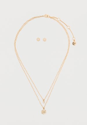 Aranyszínű, rétegezett nyaklánc, amely két láncból áll, egy hatszög alakú medállal és egy kis strasszkővel. Tartalmazza a hozzáillő hatszög fülbevalókat is.