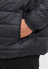 Black puffer jacket with quilted pattern, zippered pocket, and elastic cuffs, made of shiny synthetic material. Visible hand in pocket.