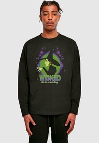 Must have pika seljae shirt, millel on mustad pikad varrukad ja millel on roheline ja lilla nõida graafika, mille all on kiri "WICKED", valmistatud pehmest puuvillasegusest materjalist.