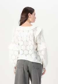 White eyelet top featuring a scalloped collar, ruffled sleeves, and cut-out floral pattern, styled with grey wide-leg pants.