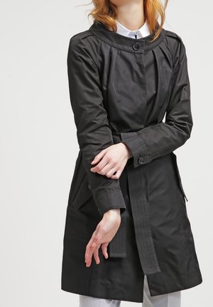 Personne portant un trench-coat noir ceinturé avec des poignets boutonnés et une chemise blanche à col en dessous, sur un fond uni.