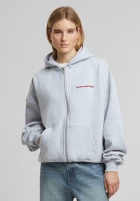 Haut à capuche gris zippé avec une poche avant, comportant le texte rouge "INTERDIT" sur la zone supérieure gauche de la poitrine et des manches à coupe décontractée.