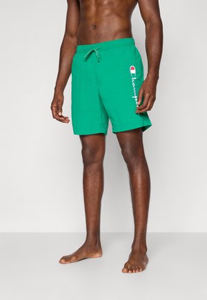 Uomo dalla pelle scura che indossa pantaloncini da bagno verdi Champion con coulisse, in piedi a piedi nudi davanti a uno sfondo chiaro e uniforme.