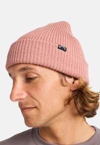 Beanie rosa a coste con bordo pieghevole e una piccola etichetta nera del brand. Indossata con abbigliamento casual, mostrando dettagli di texture e design.