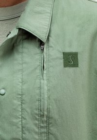 Hellgrüne Jacke mit glatter Textur, ausgestattet mit einem Reißverschluss, einem umgeschlagenen Kragen und einem gestickten Quadrat-Logo in dunklerem Grün.