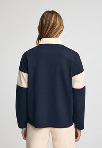 Giacca a blocchi di colore blu navy e crema con colletto, caratterizzata da una texture morbida e cuciture rosse a contrasto lungo le maniche.