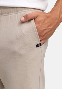 Mano derecha en el bolsillo de un pantalón beige con etiqueta de marca de tela negra visible, parte de la camisa blanca por encima de la cintura.