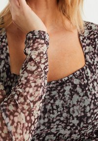 Genomskinlig blus med långa ärmar som har ett blommönster i nyanser av svart, rosa och grönt, med fyrkantig halsringning och volangdetaljer på ärmsluten.