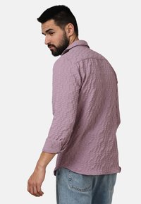 Camicia di un morbido viola chiaro con texture, colletto e maniche arrotolate, caratterizzata da un motivo decorativo. Il tessuto appare morbido e casual.