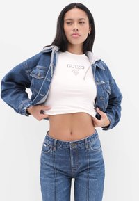 Kurtka dżinsowa z szarą podszewką bluzy z kapturem, jasnoniebieska, o pudełkowym kroju. Biały krótki t-shirt z logo "GUESS", niebieskie, wysokie dżinsy z przeszyciami.