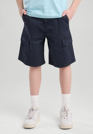 Dreng iført marineblå cargoshorts, lyseblå t-shirt, hvide sokker og hvide sneakers, hænderne i lommerne, stående på gråt gulv.