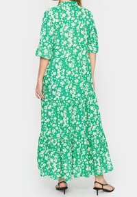 Maxiklänning i grön tyg med vit blommönster, korta uppvikta ärmar, lagerdesign och nederkant under vristerna. Bärs med svarta sandaler med remmar.