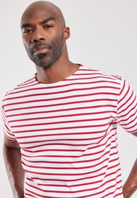 Camiseta blanca con rayas rojas horizontales, de mangas cortas y cuello redondo. La tela parece suave con un diseño ajustado.