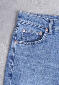 Κοντινή λήψη τσέπης από μπλε τζιν με χαλκομανίες και ορατή ραφή σε γκρι επιφάνεια τσιμέντου.