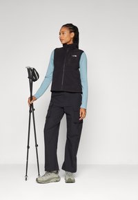Black fleece vest over a light blue long-sleeve shirt, cargo pants, and gray hiking shoes; person holds trekking poles.